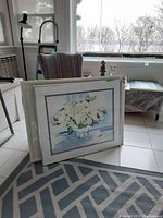 Two framed watercolours leaning against a chair near a window. One is smaller, visible on front, with white flowers in a vase on a light blue background. Second artwork is partially visible behind, similar subject and frame.