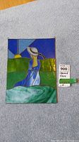Stained glass panel of woman in hat and blue dress with green hills and sun background, shown laying on a gray towel on wooden surface.