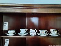 Four china teacups each with a matching saucer, displayed on a wood shelf. Also includes a small bowl and a creamer.