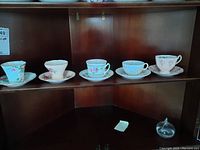 Five china teacups with their matching saucers displayed on a wooden shelf, each featuring different floral designs and colors including blue, pink, and rose patterns.