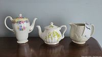 Three items on a wooden table: tall floral bone china teapot, white Staffordshire teapot with green floral design, and an off-white pottery creamer.