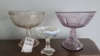 Three vintage pressed glass compotes: two large purple tinted and one smaller with milk glass trim.