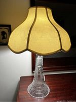 Table lamp illuminated on a wooden surface, showing the glass etched base and textured fabric scalloped shade with decorative trim.