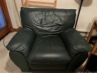Black leather oversized armchair in an indoor setting. Shows some wear and creasing on leather, wide armrests, and cushioned seat and back.