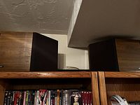 Pair of Bose wooden speakers shown placed side by side on furniture shelf. Each speaker has a black fabric grill on the front and a wooden exterior.