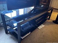 Black metal frame TV stand with multiple glass shelves, placed on carpeted floor with TV on top