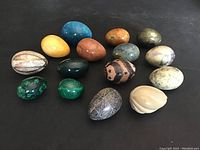 Fifteen assorted stone eggs arranged on black surface, showing variety of colors and materials.