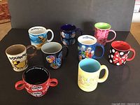 Photo showing nine assorted Disney and Universal Studios mugs arranged on a surface, featuring Mickey Mouse, Minnie Mouse, and other Disney characters.