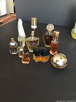 Nine assorted used perfume bottles arranged on black surface in front of neutral background. Various colors and shapes including bottles with gold caps, black caps, and decorative stoppers.