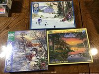 Photo showing three factory sealed 1000-piece jigsaw puzzles with winter and summer scenes on the box covers.
