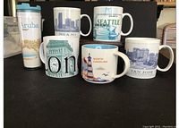 Seven items including six city-themed ceramic Starbucks mugs and one tall travel tumbler with Aruba design shown on black background.