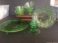 Photo showing seven green uranium glass pieces including cake plate, candle holder, and two matching bowls with ribbed pattern.