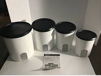 Four white Tupperware canisters with black one-touch lids arranged from largest to smallest with a small instruction booklet in front.