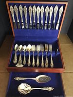 Photo showing forks on the upper section of the case with silverplate ornate handles and teaspoons and forks below in blue velvet slots. Two silver serving utensils lay in front of the lower section.