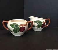 Two ceramic cream and sugar pieces with apple and leaf design, handles look like branches, shown from side on black background