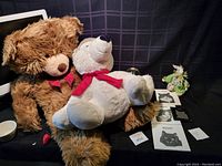 Two plush bears, one brown with red bow and one white with red scarf, sitting on a dark surface with some small decorative items and cat coasters.