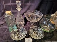 Glass fishbowl, two patterned candy bowls, brandy decanter displayed together on black cloth