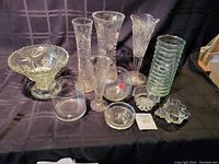Wide view of the lot items arranged on a black surface, showing various sizes of glass vases, bowls, and a stack of glass plates.