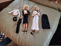 Two vintage Barbies laid out with accompanying accessories like jackets, hats, stockings, skirt, and shoes on sofa.