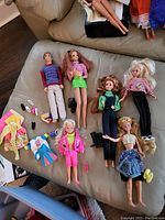Six vintage dolls including Barbie, Ken, Skipper, Maxi and various outfits and shoes spread on a beige couch.
