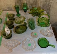 Overview of all 15 glass items on display showing variety of green and clear glass bowls, vases, a lidded chicken dish, a green glass basket and a saucepan style dish.