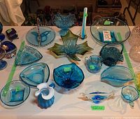 Overview of 15 turquoise blue glass items including bowls, plates, candy dishes, and art glass on a white table with green tape for sale display.