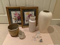 Photo of two framed floral prints, three ceramic vases, one glass candle holder, and three decorative glass birds arranged on a pink cloth surface.