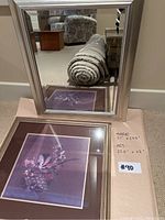 Silver framed mirror standing behind the floral art print on floor.