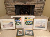 Full view of five framed artworks positioned in front of a stone fireplace. Three larger pieces behind, two smaller on floor in front.