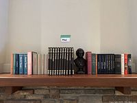 Wide view of the collection of hardcover books and the Mozart bust sculpture on a wooden shelf above a stone wall.
