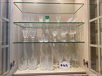 Full cabinet view of 16 assorted crystal glasses showcasing various sizes and patterns on three shelves.