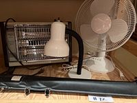 Photo showing electric heater, desk lamp and oscillating fan on shelf with pool cue case in front