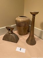 Photo of three decor pieces on a beige surface against a tan wall: a gold metal candlestick holder, a decorative jar with lid, and a small decorative vase.