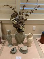 Front view showing six ceramic vases with artificial dried flowers in largest vase. Vases in muted beige and greenish tones, some with carved or ribbed designs.