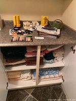 Countertop and cabinet shelves with folded linens and various miscellaneous household items including Eveready flashlight, mugs, watering can, thread spools, and small tools.