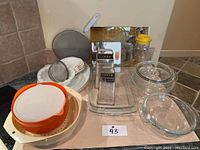 Photo showing various kitchenware items on a counter including bowls, strainers, glass jars, plates, a box grater, and glass baking dishes.