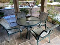 Full view of round glass top table surrounded by 4 matching metal frame chairs with patterned fabric seats and backs on stone patio under covered porch.