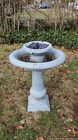 Full view of weathered bird bath fountain standing on grass with visible brown leaf debris, showing basin and central fountain area.