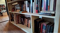 Wide view of bookshelves containing a mix of hardcover and paperback novels, some titles visible, books organized upright and stacked horizontally in some places.