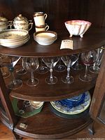 Curved corner cabinet with two shelves displaying glass stemware and porcelain dishes including covered pots, bowls, and plates with floral patterns and gold accents