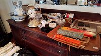 Wide view of the dresser with anniversary clock under glass dome, Swarovski crystal clock, blue transferware pitcher and bowl, collector's plates stacked, ashtrays, lighters, chopsticks in case, and the large silver serving tray on the right.