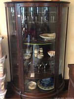 Front view of vintage wooden curio cabinet displaying various glassware and ceramic dishes inside, showing curved glass panels and wooden frame.