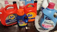 Multiple laundry supplies including two large orange Tide detergent jugs, one small blue Clorox detergent bottle, Downy fabric softener jug, and bleach bottle on dark surface against stone wall.