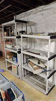 Two assembled gray plastic shelving units against a white stone wall in a basement. One unit contains various stored items.