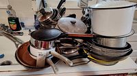 Overview of assorted pots and pans stacked in kitchen sink area, shows metal and enamel cookware in various sizes with many handles and lids.