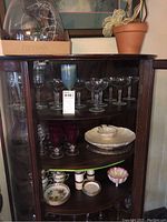 Glass cabinet showing multiple shelves with clear coupe and champagne glasses, red stemware, Lenox shrimp cocktail platter, and small porcelain dishes.