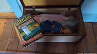 Opened wooden toy box on wooden floor showing doll(s) and stacked books inside