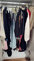 Collection of women's clothing hanging inside closet, includes various jackets, blouses, dresses in dark and light colors.