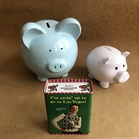 Front view of three coin banks: large blue ceramic piggy bank, smaller pink CIBC plastic piggy bank, and rectangular Las Vegas savings tin.