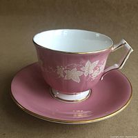 Side angle of footed dusty-rose Aynsley cup on matching saucer showing grapevine motif and gold trim.